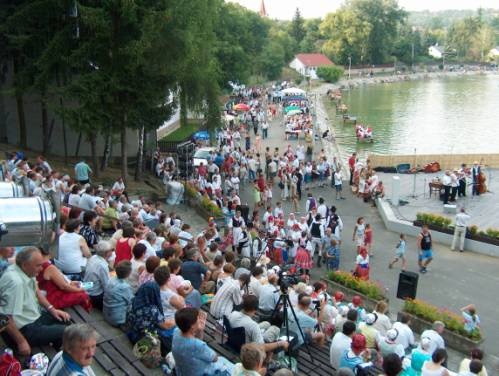 A Vízi Színpad lelátója látható, emberek ülnek a padokon. A háttérben a tó partján sétáló tömeg látható és a színpad kis része is belóg a képbe.