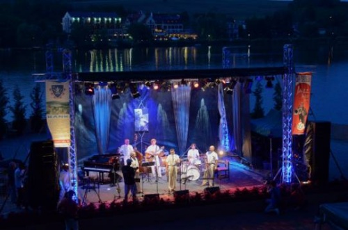 Koncert a Vízi Színpadon. Este van, a reflektorok fénye bevilágítja a színpadot. A közönség soraiban sötét van, a háttérben fények csillognak a tavon.