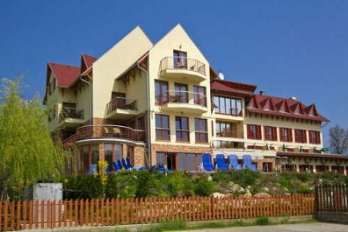 A hotel többszintes épülete látható a képen. A sárga épület teteje háromszög formájú csúcsban végződik. Sok üvegfelület, sok ablak látható rajta. A földszinti teraszon kék napozószékek sorakoznak.