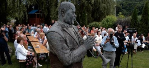 A szobor hátterében a szabadtérben kihejezett asztalonál néhány tucat ember ül.